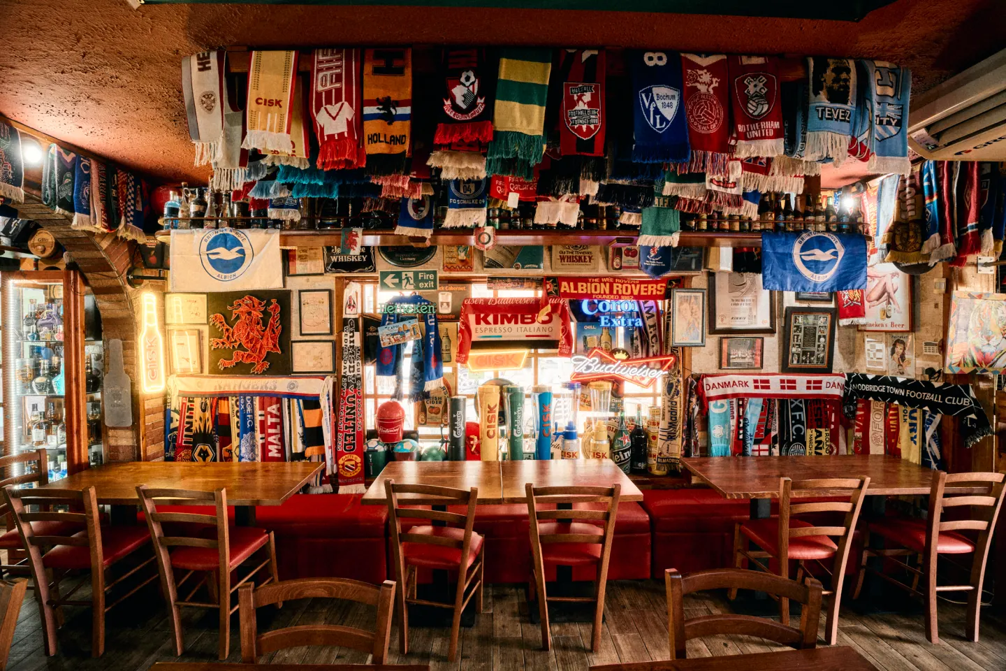 The back room: tables and red benches under a ceiling of football scarves, the taps and neon signs behind