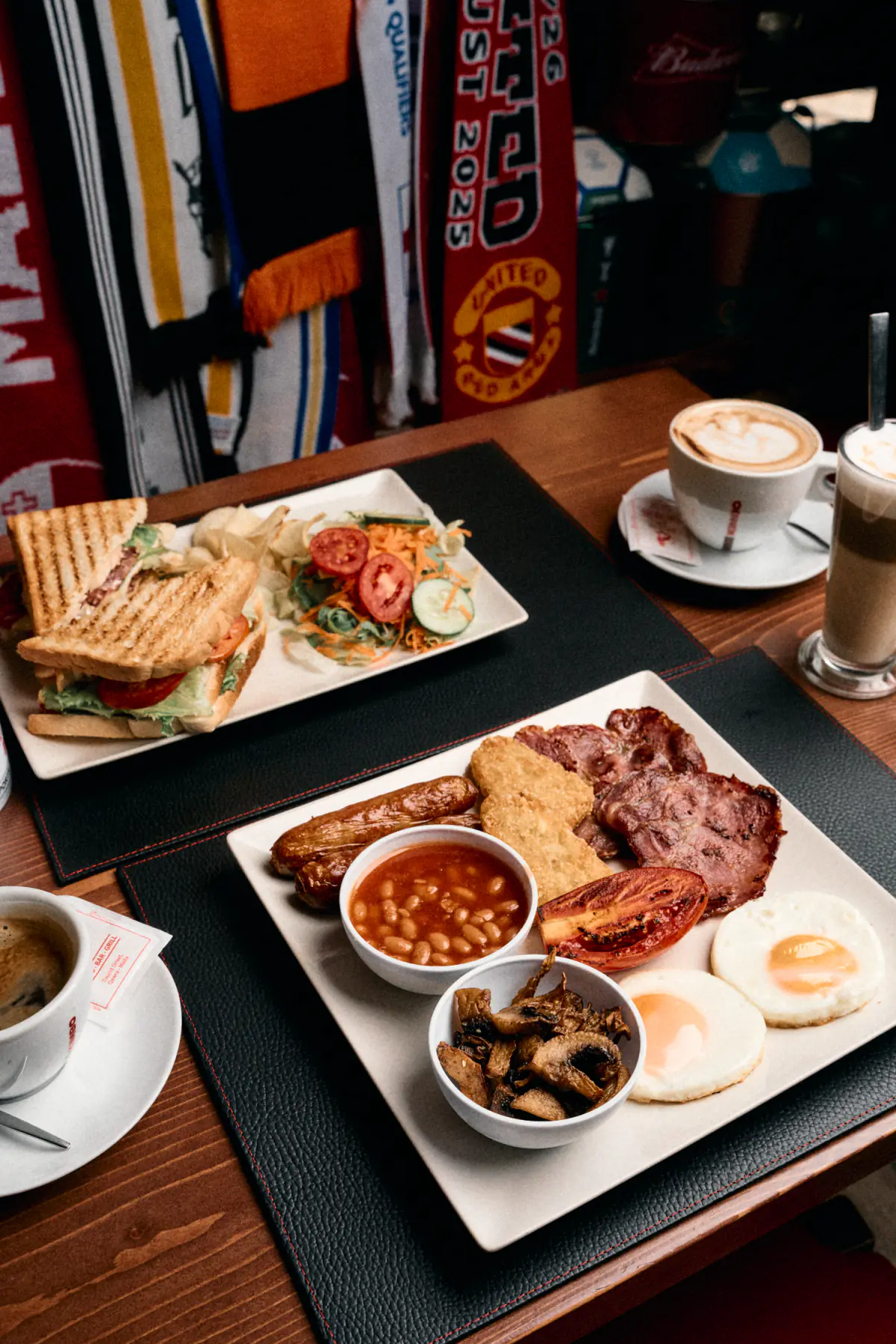 A full breakfast of eggs, bacon, sausages, beans, mushrooms and hash browns, with a toasted sandwich and coffees behind